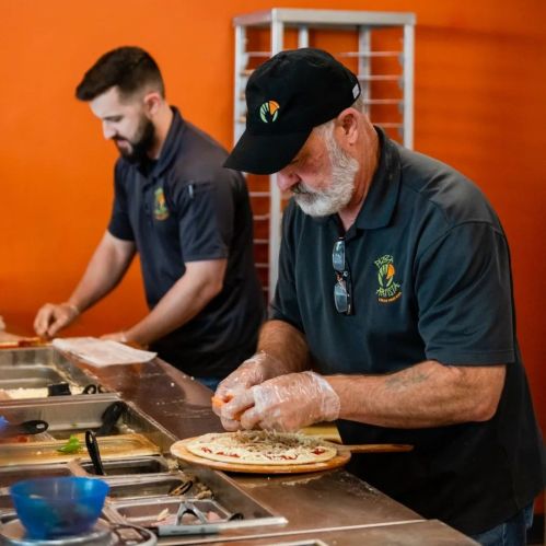 Two men prepare pizzas in a kitchen, one chopping ingredients and the other working in the background.