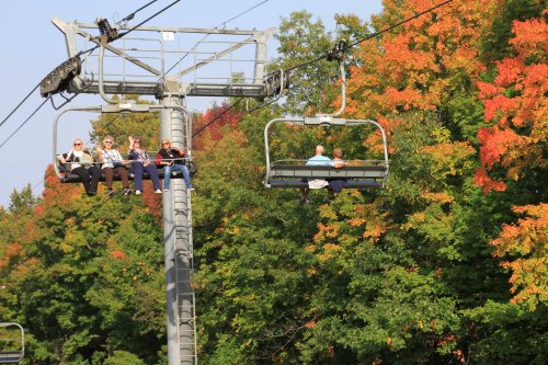 Two ski lift chairs with people riding above vibrant autumn foliage.