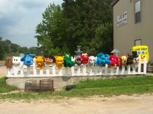 A row of colorful animal heads mounted on a white fence, with a cartoon character head at the end, set in a rural area.