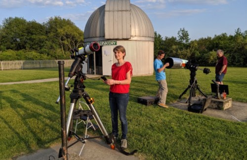 A woman stands by a telescope outside an observatory, while another person sets up a telescope nearby on a grassy field.