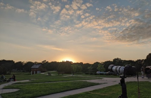 A serene park scene at sunset, featuring a camera on a tripod and a colorful sky with scattered clouds.