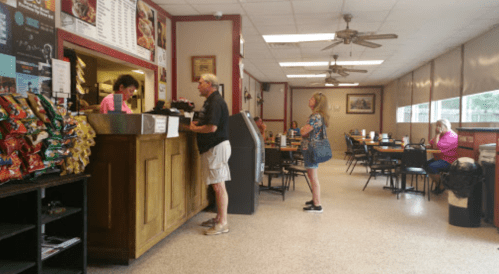 A casual restaurant interior with customers at the counter, tables, and snacks displayed on a shelf.