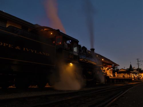 A steam locomotive at dusk, releasing smoke near a vintage train station illuminated by warm lights.