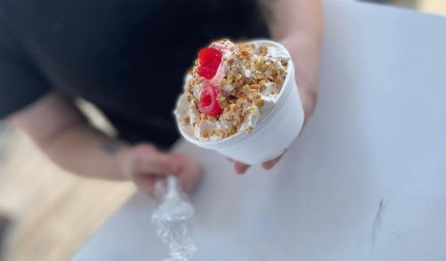 A person holds a cup of dessert topped with whipped cream, popcorn, and raspberries.