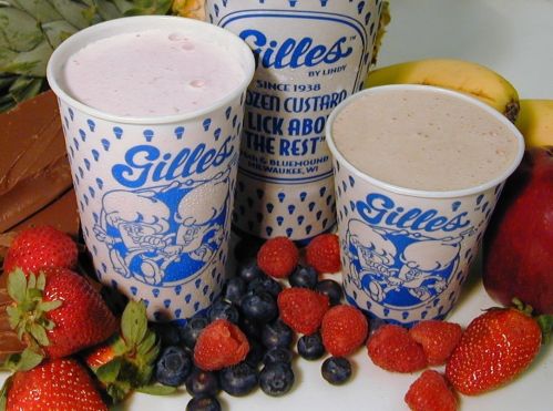 Two cups of frozen custard in blue and white cups, surrounded by fresh strawberries, blueberries, bananas, and an apple.