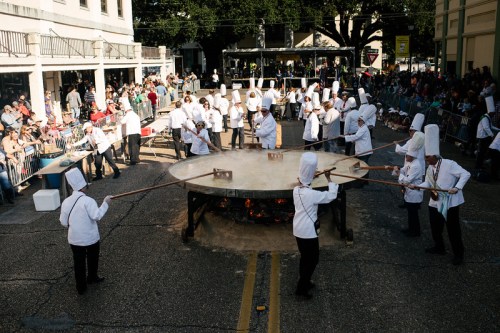 A large outdoor cooking event with chefs in white hats stirring a massive pot over an open flame, surrounded by spectators.