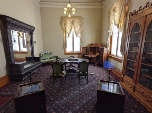 A vintage room featuring a table, chairs, a sofa, and a bookshelf, with elegant curtains and decorative lighting.
