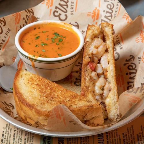 A plate with a grilled shrimp sandwich and a bowl of tomato soup, garnished with herbs.
