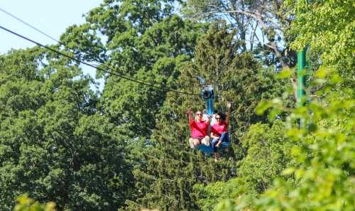 Two people in red shirts ride a chairlift through a lush green landscape, waving and enjoying the view.