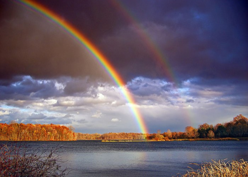 A vibrant double rainbow arcs over a tranquil lake surrounded by autumn trees under a dramatic sky.