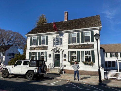 A white historic building with green shutters, a flag, and a sign for "Goods & Curiosities," with a man standing nearby.