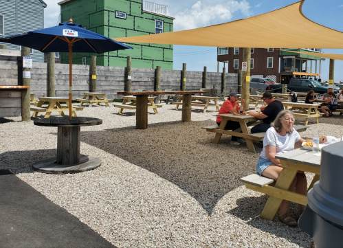 Outdoor dining area with picnic tables, shade sails, and patrons enjoying food in a gravel setting.