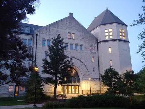 Historic stone building with large archway, illuminated windows, and surrounding trees at dusk.
