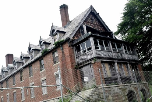 A large, dilapidated mansion with a slanted roof, overgrown vegetation, and a chain-link fence surrounding it.