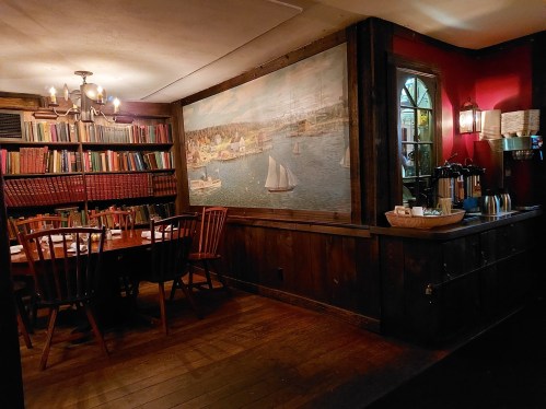 Cozy dining area with wooden furniture, a large mural of a waterfront, and shelves filled with books.