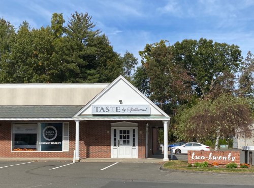 Exterior of "Taste by Gelato Mund," a gourmet bakery, with a sign for "two-twenty" nearby and trees in the background.