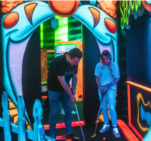 Two people play mini-golf in a colorful, neon-lit indoor course with a clown-themed entrance.