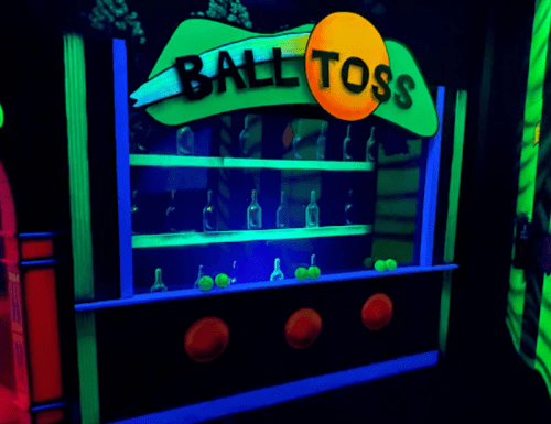 A brightly lit arcade game called "Ball Toss" with bottles on shelves and three red buttons in front.