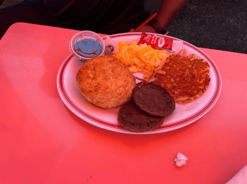 A plate of breakfast food featuring a biscuit, scrambled eggs, hash browns, sausage patties, and condiments.