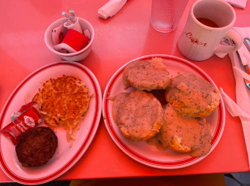 A plate of biscuits smothered in gravy, hash browns, a sausage patty, and a cup of coffee on a red table.
