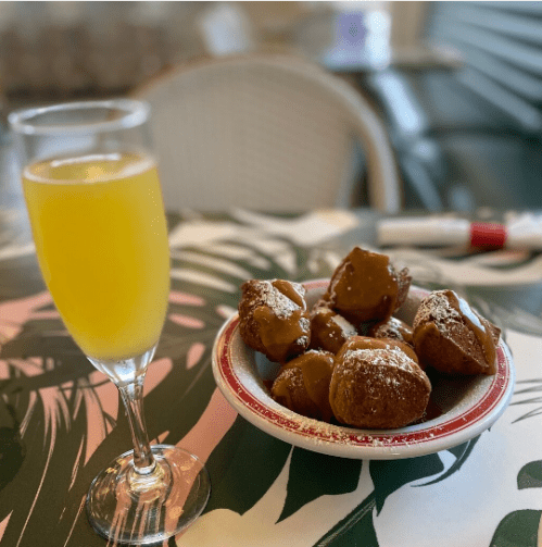 A glass of mimosa beside a plate of golden-brown pastries drizzled with caramel on a tropical-themed table.