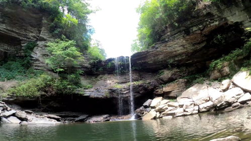 A serene waterfall cascades into a calm pool, surrounded by lush greenery and rocky cliffs.