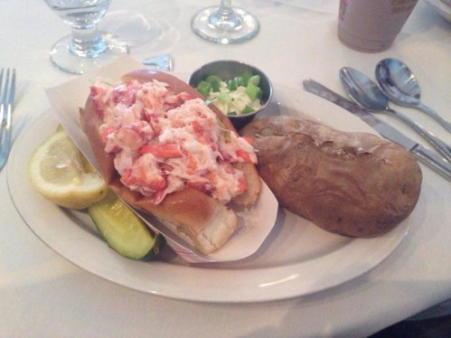 A lobster roll topped with lobster salad, served with a baked potato, lemon wedge, and pickles on a white plate.