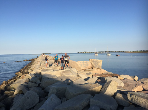 A group of people walking on a rocky jetty by the calm sea, with boats in the distance and clear blue skies.