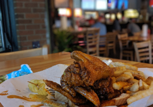 A plate of fried chicken stacked on a bun, served with crinkle-cut fries and pickles in a casual dining setting.