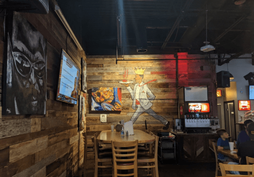 Interior of a restaurant featuring wooden walls, artwork, a TV, and tables with chairs. Warm lighting creates a cozy atmosphere.