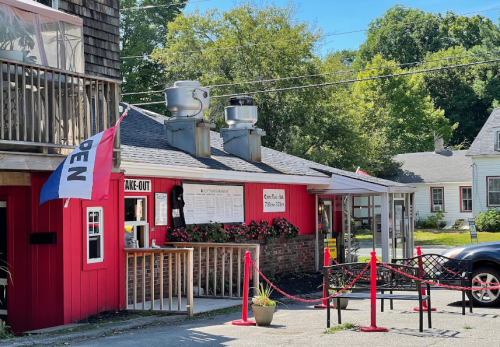 A red take-out restaurant with a menu board, outdoor seating, and a flag indicating it's open. Trees in the background.