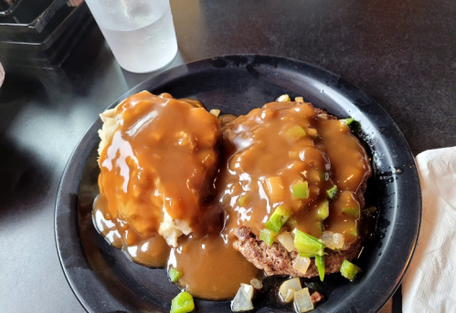 A black plate with two meat patties covered in brown gravy, served with mashed potatoes and green bell pepper pieces.