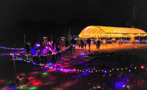 A nighttime scene with colorful lights illuminating a path leading to a tent filled with people.
