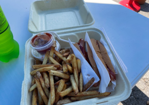A takeout container with two hot dogs, fries, and a small cup of ketchup, alongside a green soda.
