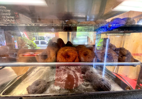 A display case filled with various donuts, some dusted with powdered sugar, viewed through glass.