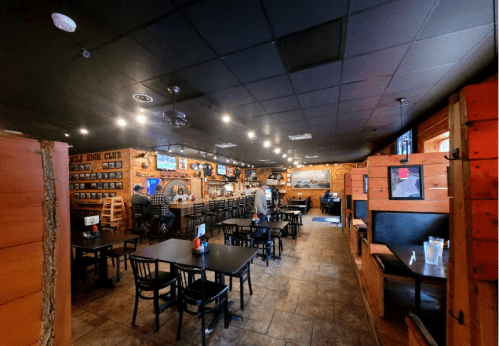 Interior of a rustic bar with wooden walls, tables, and a long counter, featuring TVs and a cozy atmosphere.
