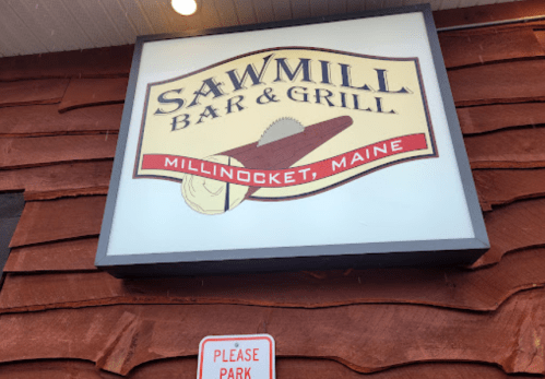 Sign for Sawmill Bar & Grill in Millinocket, Maine, featuring a saw and wood design. "Please park" sign below.