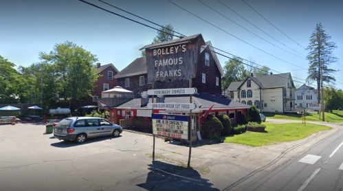 A roadside eatery named "Bolley's Famous Franks" with a sign and outdoor seating, surrounded by trees and houses.