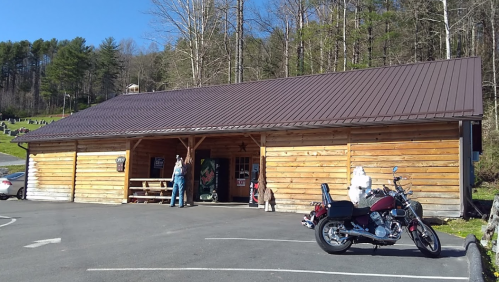 A wooden building with a metal roof, parked motorcycles, and people outside in a rural setting surrounded by trees.