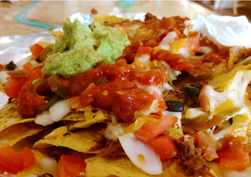 A close-up of nachos topped with cheese, salsa, guacamole, diced tomatoes, and shredded meat on a foil tray.