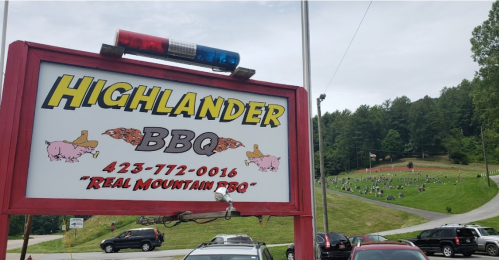 Sign for Highlander BBQ with a police light on top, set against a backdrop of a grassy area with gravestones.
