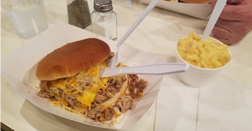 A pulled pork sandwich topped with cheese, served with a side of macaroni and cheese, on a table with condiments.