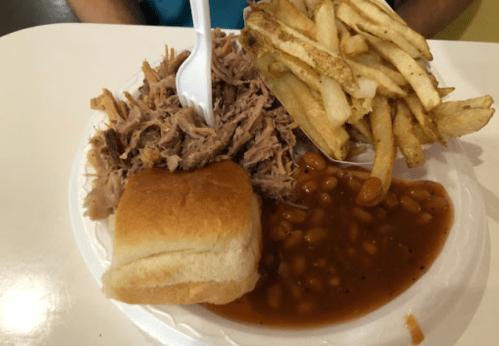 A plate with pulled pork, baked beans, a roll, and French fries on a white paper plate.