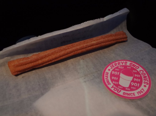 A single churro on parchment paper next to a circular sticker for Jerry's Sno Cones.