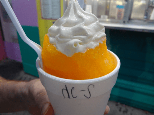 A hand holding a cup of orange snow cone topped with white whipped cream.