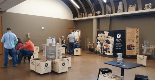 A spacious room with people engaging in activities, surrounded by wooden structures and KEVA blocks on display.