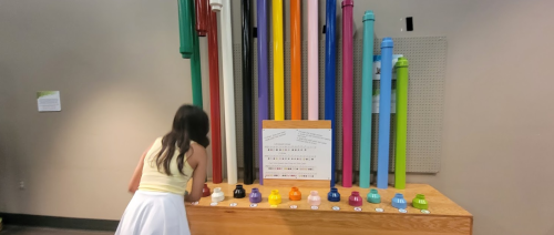 A person examines colorful pipes displayed vertically on a wooden table, with a sign and buttons in front.