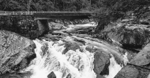 A black and white image of a rushing river flowing under a stone bridge, surrounded by rocky terrain and trees.