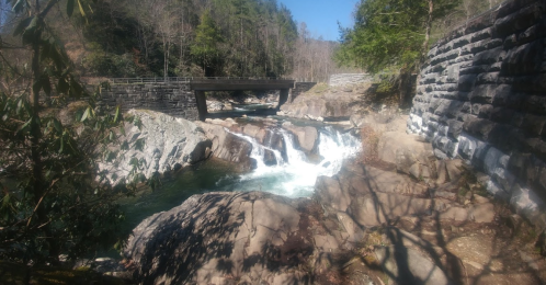 A serene waterfall cascades over rocks beneath a bridge, surrounded by trees and a rocky landscape.