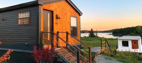 A cozy tiny house with a wooden exterior, surrounded by greenery and a serene lake at sunset.